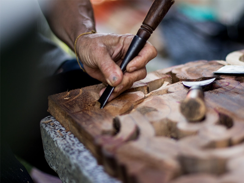 Close-up of artisan hands using chisel to hand-carve intricate patterns into reclaimed teak wood