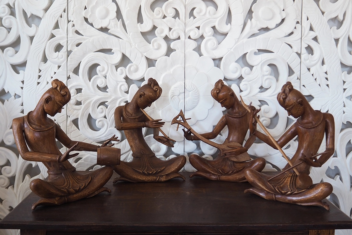Handcrafted wooden musician sculptures from Thailand displayed in front of a white carved teak backdrop.
